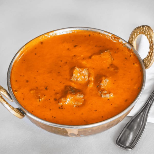 A copper bowl filled with orange curry, containing paneer cubes, sits on a white tablecloth with a spoon.
