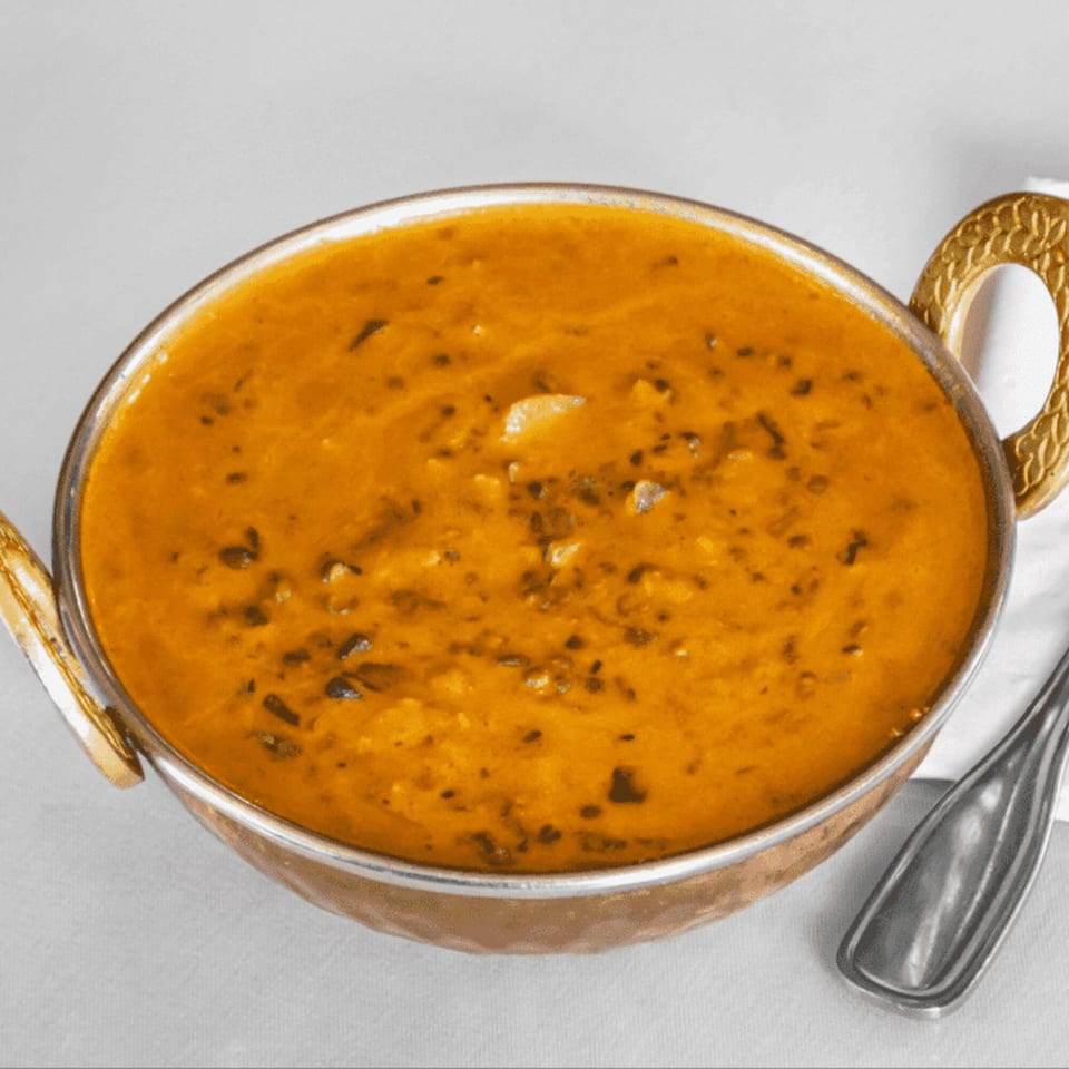 A copper bowl filled with orange lentil dal, garnished with herbs, next to a spoon.