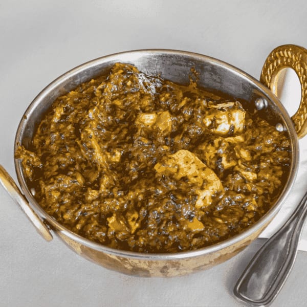 A copper bowl filled with vibrant green palak paneer, featuring chunks of cheese in a rich spinach sauce.